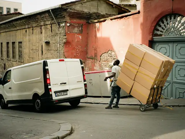 Worker wheeling a tall stack of boxes on a hand truck toward a white van