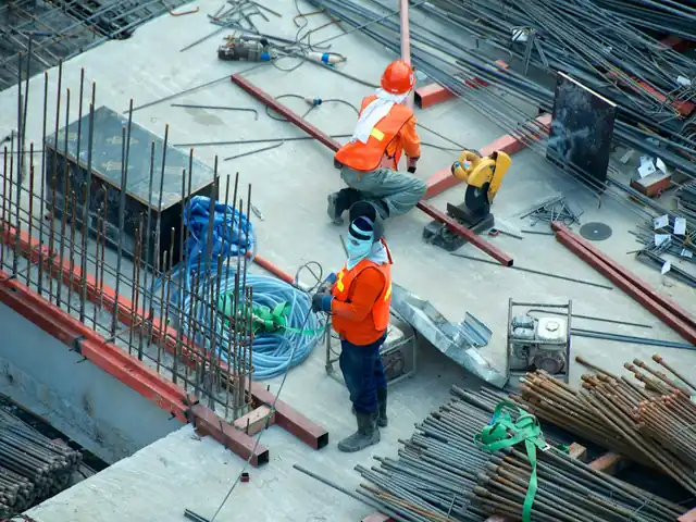 Two workers in orange vests on a concrete deck among steel bars and beams